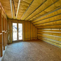 Poddasze domu w trakcie ocieplania: wełna mineralna między metalowymi profilami, okno z widokiem na domy, surowa podłoga z płyt OSB.