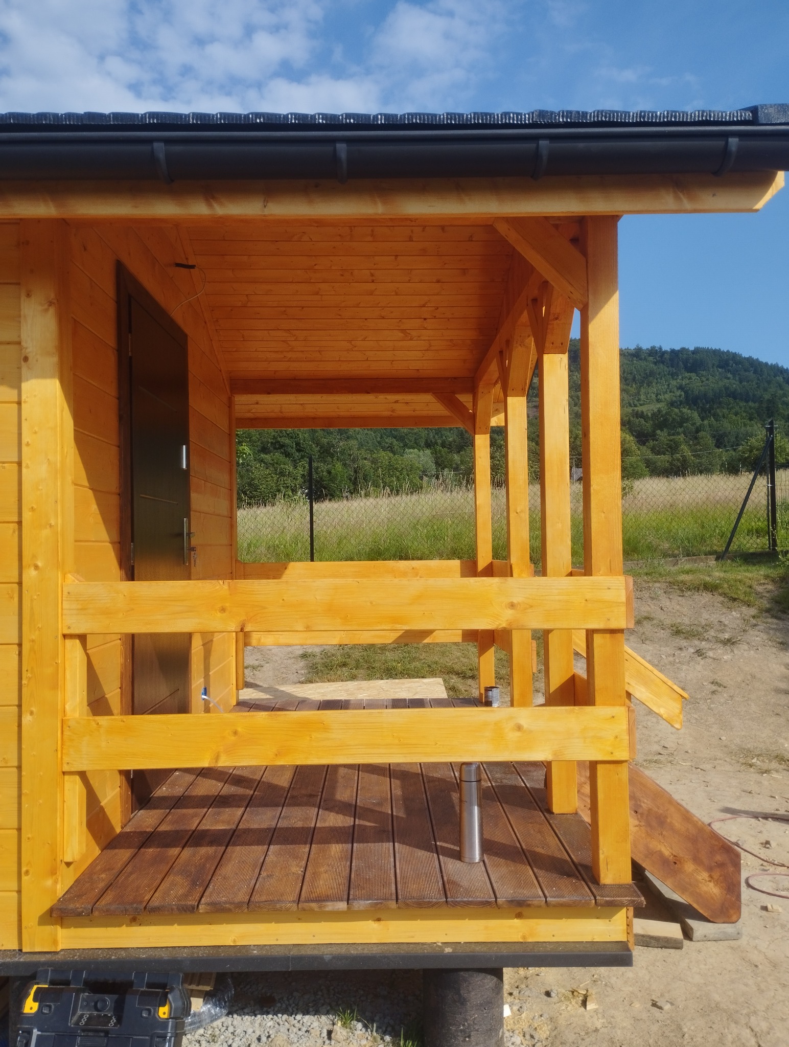 Drewniana altana z tarasem na tle zieleni. Widoczna konstrukcja z belek, podłoga z desek i metalowy termos na tarasie. Całość w jasnym, naturalnym odcieniu drewna.