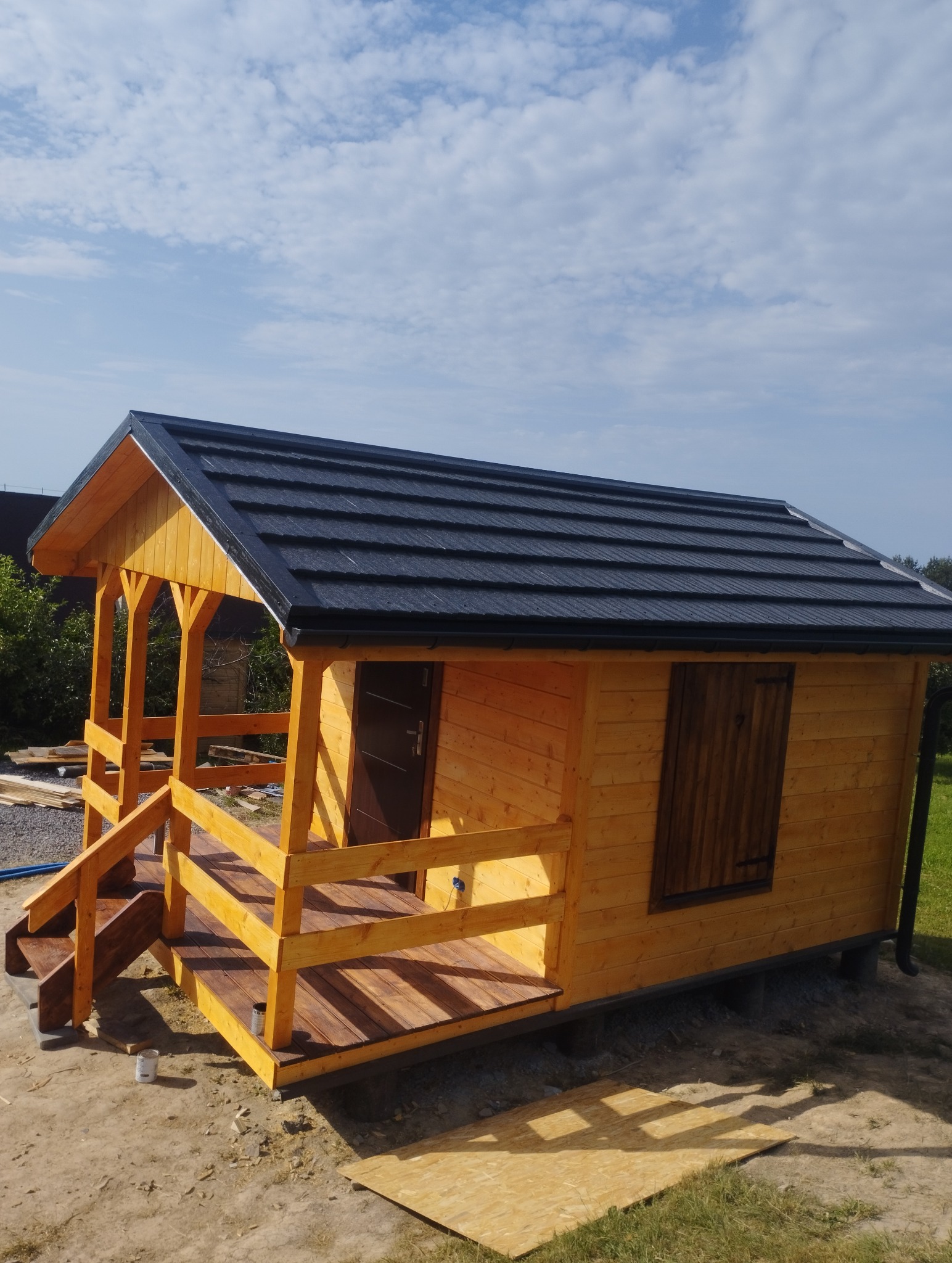 Drewniany domek letniskowy z zadaszonym tarasem i schodami, wykończony w jasnym odcieniu drewna, z ciemnym dachem z blachodachówki. Błękitne niebo w tle.