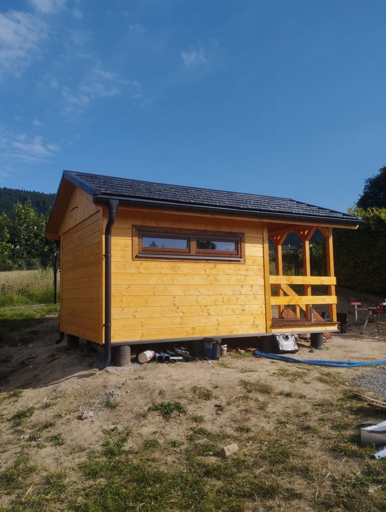 Żółty, drewniany domek narzędziowy z czarną, spadzistą dachówką, z małym oknem i zadaszonym wejściem, na tle błękitnego nieba i zieleni. Widoczne narzędzia.