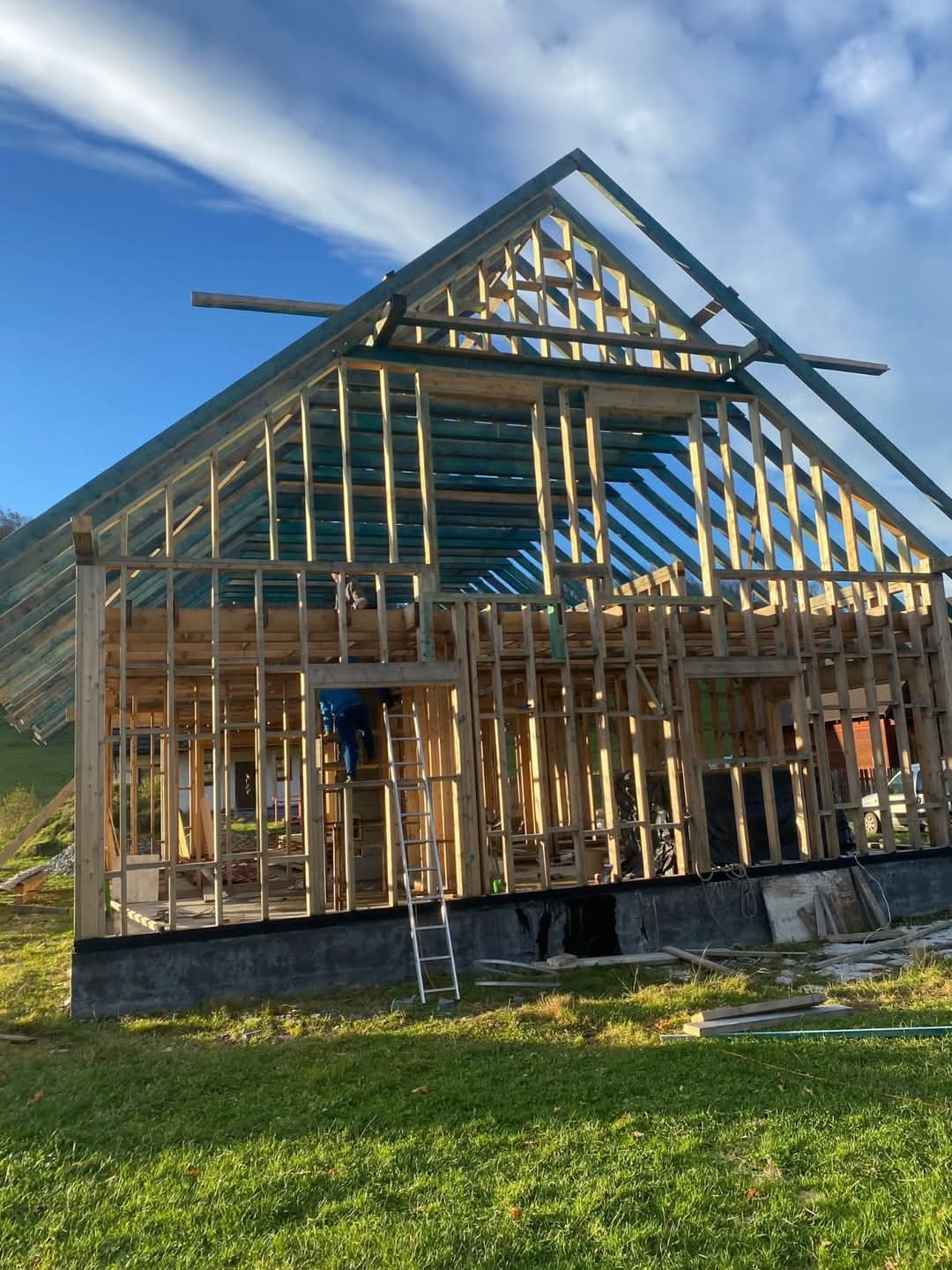 Wznoszenie drewnianej konstrukcji domu szkieletowego w słoneczny dzień. Widoczne elementy szkieletu, drabina i pracownik w niebieskim kombinezonie.