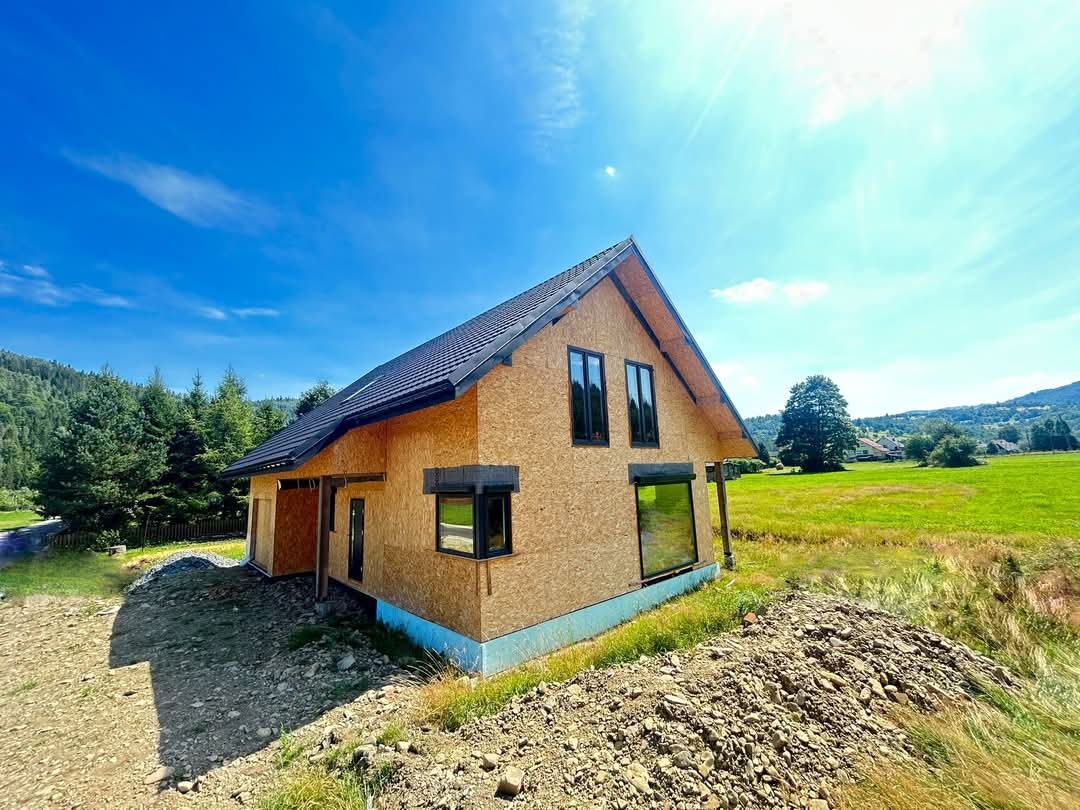 Dom szkieletowy w Rycerce Dolnej w trakcie budowy, z czarną dachówką, okna z czarnymi ramami, na tle zieleni gór i błękitnego nieba.