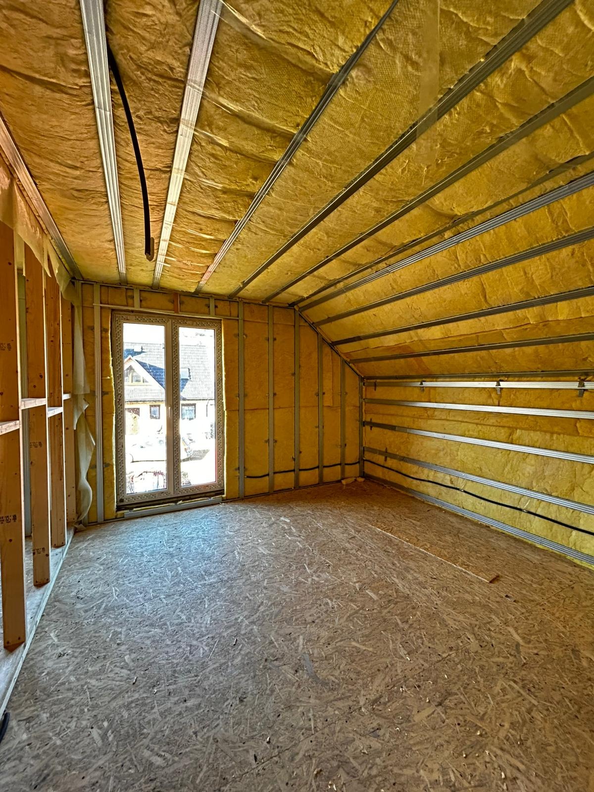 Poddasze domu w trakcie ocieplania: wełna mineralna między metalowymi profilami, okno z widokiem na domy, surowa podłoga z płyt OSB.