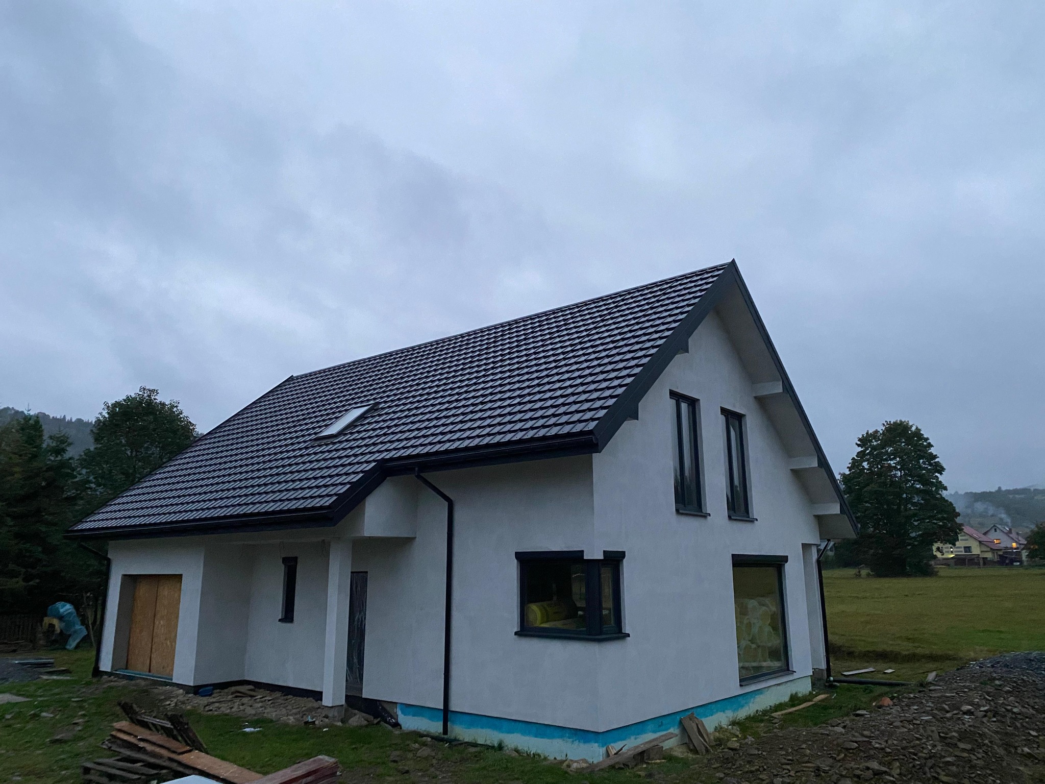 Biały dom jednorodzinny z ciemną dachówką i stolarką okienną, w tle zielone wzgórza i pochmurne niebo. Część domu jest jeszcze w stanie surowym.