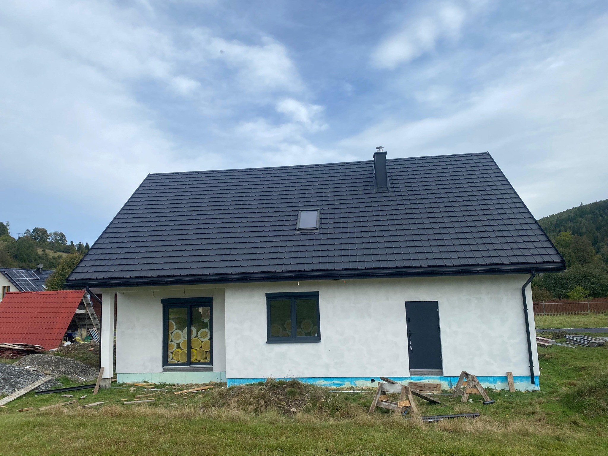 Nowoczesny dom szkieletowy z czarnym dachem i oknami w trakcie wykończenia elewacji, widok z zewnątrz na tle zieleni i pochmurnego nieba.