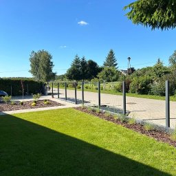 Michał Piotrowski - Zadbany trawnik z rolki, rabaty z korą i niskimi roślinami, ścieżka z kostki brukowej oddzielona metalowymi słupkami. Błękitne niebo w słoneczny dzień.