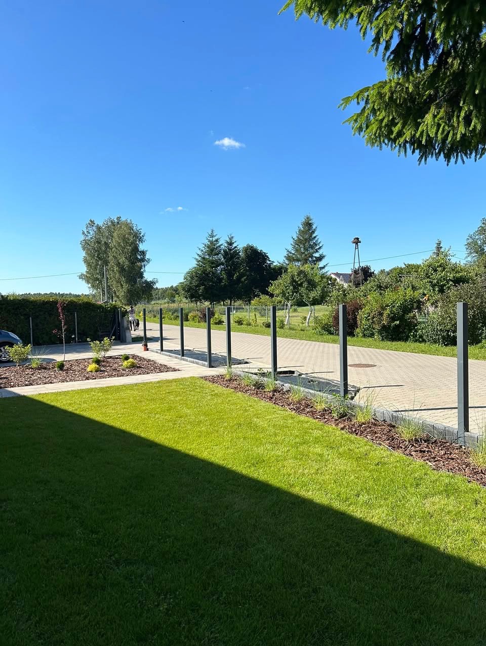 Zadbany trawnik z rolki, rabaty z korą i niskimi roślinami, ścieżka z kostki brukowej oddzielona metalowymi słupkami. Błękitne niebo w słoneczny dzień.