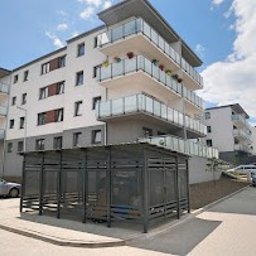 PÓŁNOC NIERUCHOMOŚCI ODDZIAŁ BOLESŁAWIEC - Nowoczesny, czteropiętrowy budynek mieszkalny z balkonami, widziany z perspektywy ulicy, z zadaszonym parkingiem rowerowym na pierwszym planie i samochodami w tle.