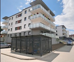 Nowoczesny, czteropiętrowy budynek mieszkalny z balkonami, widziany z perspektywy ulicy, z zadaszonym parkingiem rowerowym na pierwszym planie i samochodami w tle.