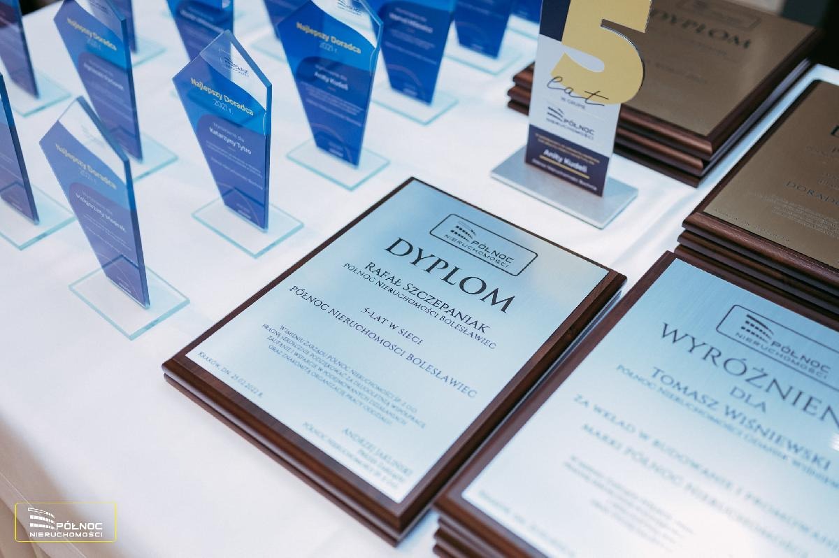 Dyplomy i nagrody 'Najlepszy Doradca' Północ Nieruchomości, w tym dyplom dla Rafała Szczepaniaka za 5 lat w sieci, na białym stole, z logo firmy. Ujęcie z góry.