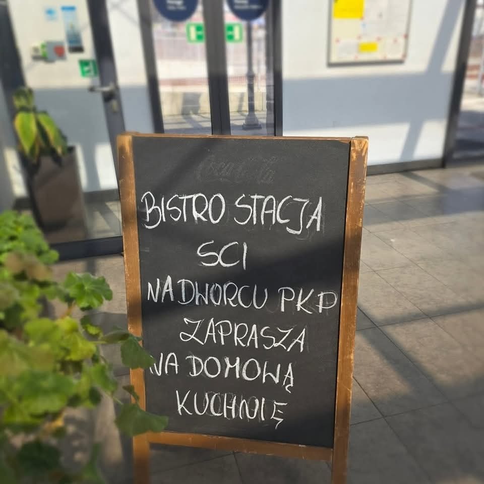Czarna tablica z menu bistro stacji SCI na dworcu PKP, zapraszająca na domową kuchnię, ustawiona przed wejściem, z zielonymi roślinami w tle.