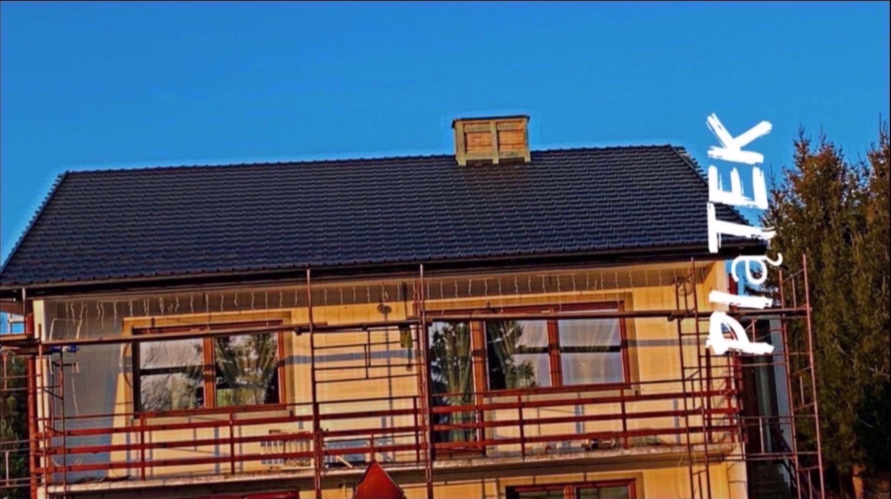 Dom w rusztowaniach z nowym, ciemnym dachem i kominem. Widoczne detale wykończenia dachu. Błękitne niebo w tle.