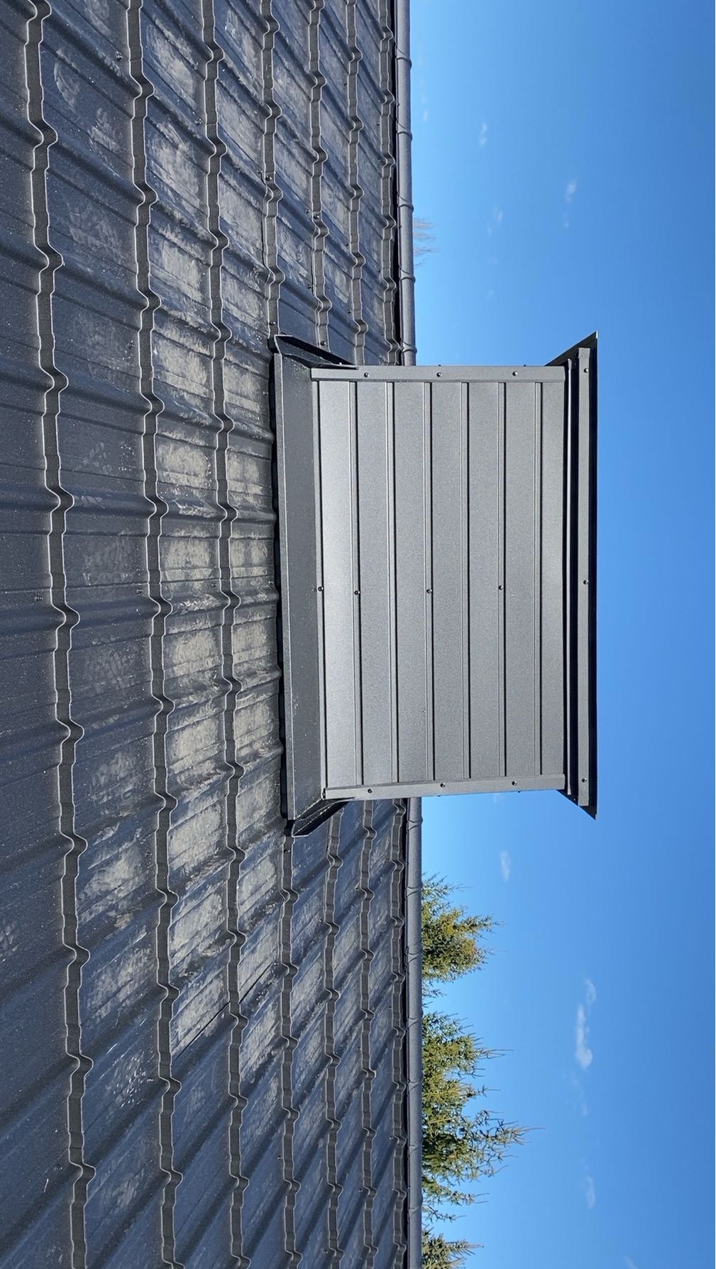 Szara blacha trapezowa na dachu z zamontowanym, metalowym wyłazem dachowym, na tle błękitnego nieba i zielonych drzew. Widok z dołu.