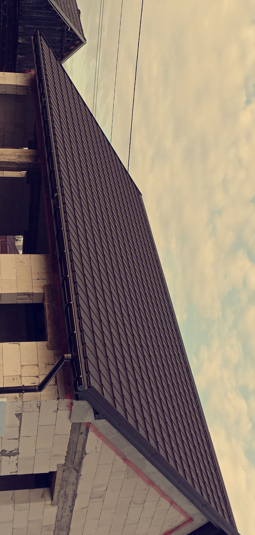 Ciemnobrązowe pokrycie dachowe z rynnami na budynku w trakcie budowy, widok z dołu, pochmurne niebo w tle. Charakterystyczny kąt ujęcia.