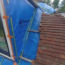 Rolldach - Prace dekarskie: fragment dachu z częściowo ułożoną dachówką ceramiczną, zabezpieczony niebieską membraną paroprzepuszczalną z logo producenta, drewniane łaty. Dach skośny.