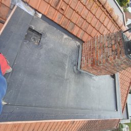 Rolldach - Widok z góry na płaski dach pokryty papą, z kominem obłożonym blachą i fragmentem dachu z dachówki ceramicznej. Prace dekarskie w toku.