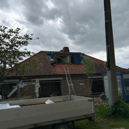 Rolldach - Demontaż uszkodzonej więźby dachowej z kominem na budynku z czerwonej cegły, widok z poziomu ulicy, z przyczepą i sprzętem budowlanym w kadrze.