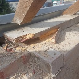 Rolldach - Fragment uszkodzonej więźby dachowej z widocznymi śladami zniszczeń, w tle zabudowa miejska. Drewno częściowo rozwarstwione, z ubytkami.