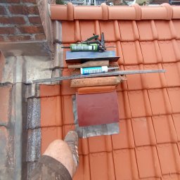 Rolldach - Czerwona dachówka ceramiczna z narzędziami dekarskimi i fragmentem nogi dekarza w ubrudzonych spodniach roboczych w rogu kadru. W tle zabudowa miejska.