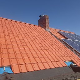 Rolldach - Czerwona dachówka ceramiczna na dachu domu, z kominem i panelami słonecznymi. Fragment dachu w trakcie układania, widoczne narzędzia i membrana dachowa.