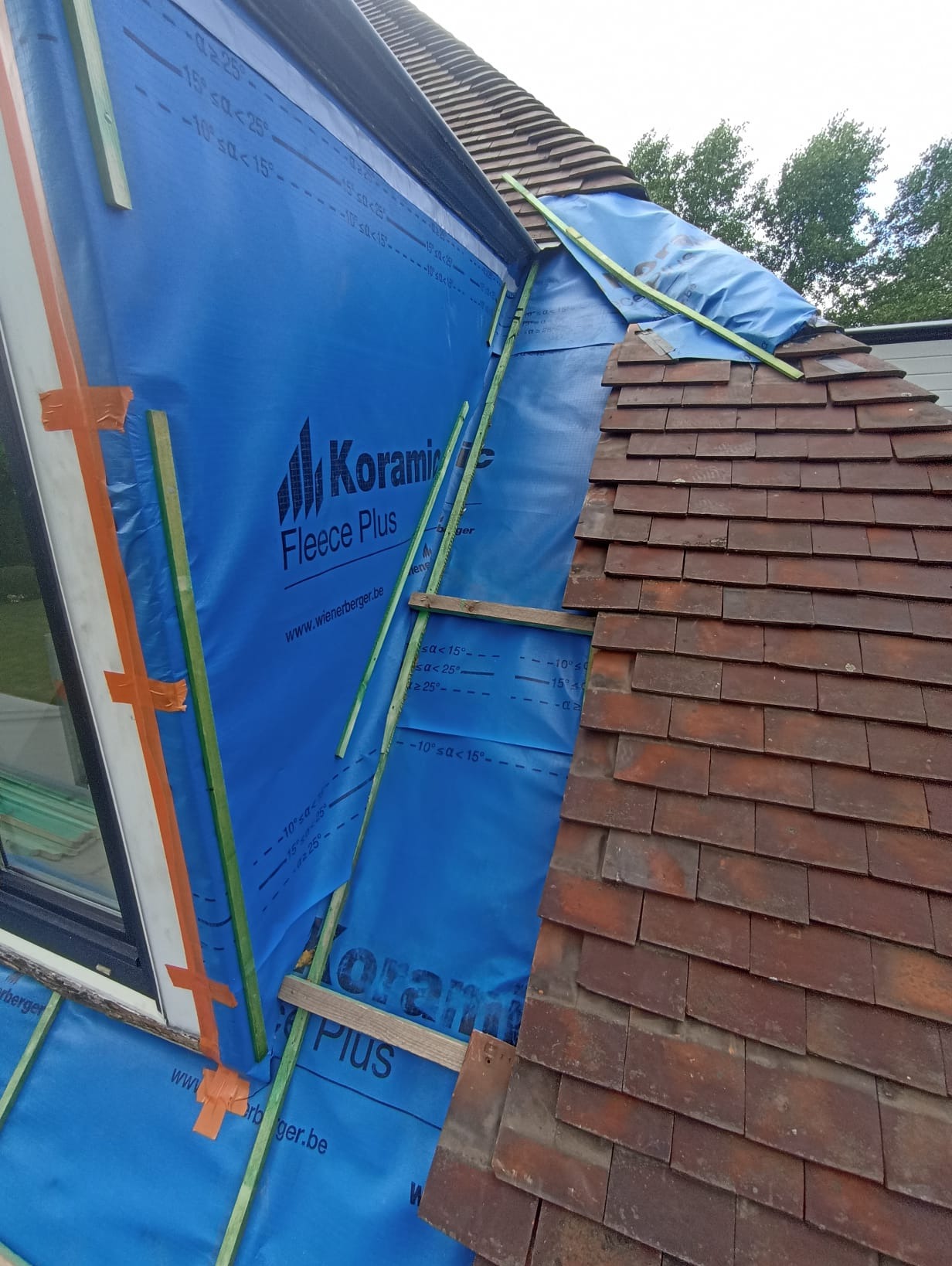 Prace dekarskie: fragment dachu z częściowo ułożoną dachówką ceramiczną, zabezpieczony niebieską membraną paroprzepuszczalną z logo producenta, drewniane łaty. Dach skośny.
