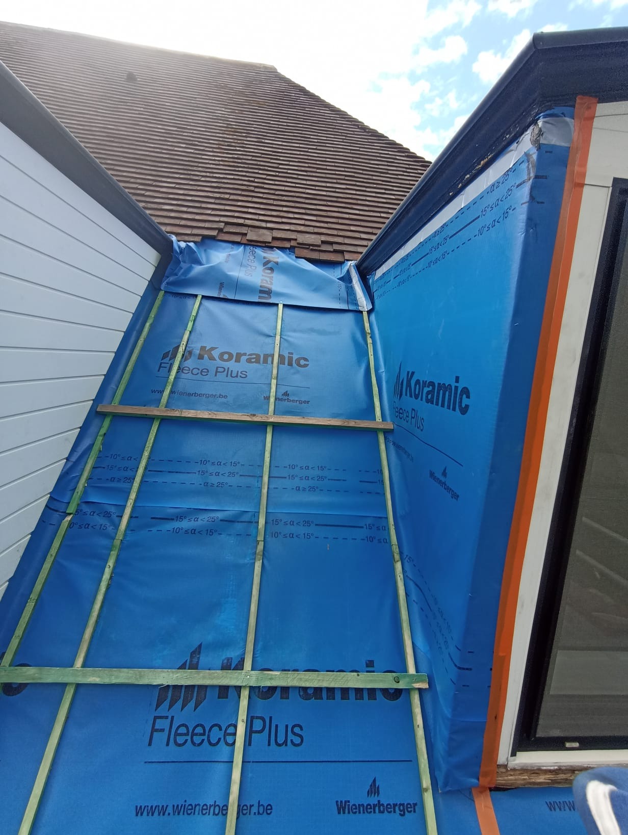 Dach z membraną w kolorze niebieskim, na niej drewniane łaty, widok z góry, fragment okna i dachówki. Prace dekarskie w toku, zabezpieczenie dachu przed wilgocią.