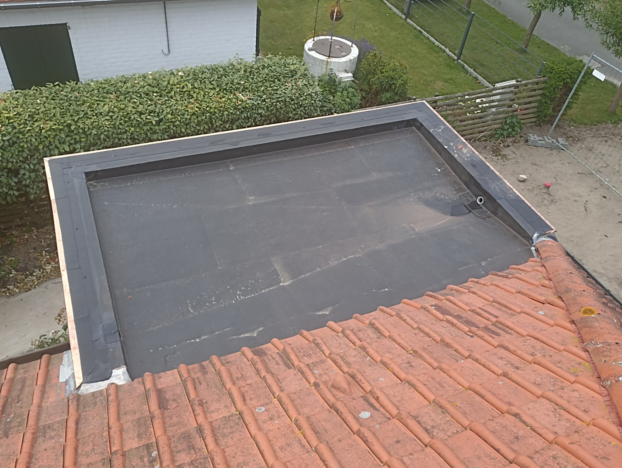 Wykończony płaski dach z czarną papą, otoczony obróbką blacharską. Widok z góry, w tle zieleń, budynek gospodarczy i studnia. Dachówka w kolorze terakoty na pierwszym planie.