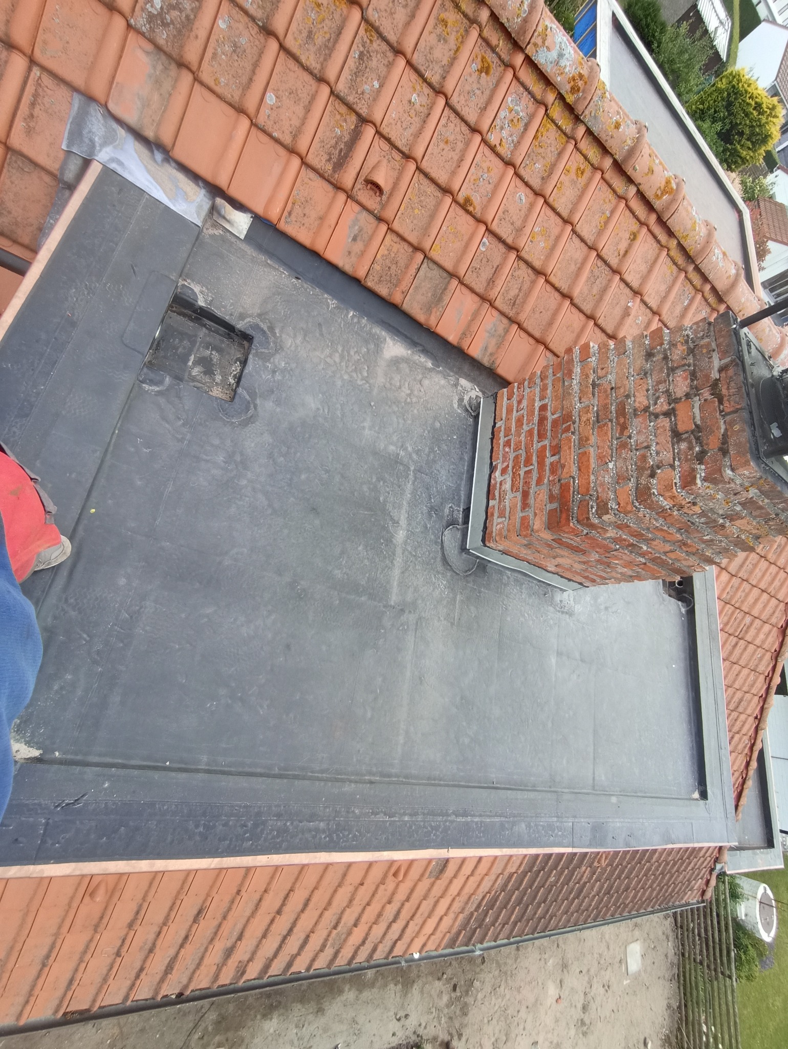 Widok z góry na płaski dach pokryty papą, z kominem obłożonym blachą i fragmentem dachu z dachówki ceramicznej. Prace dekarskie w toku.