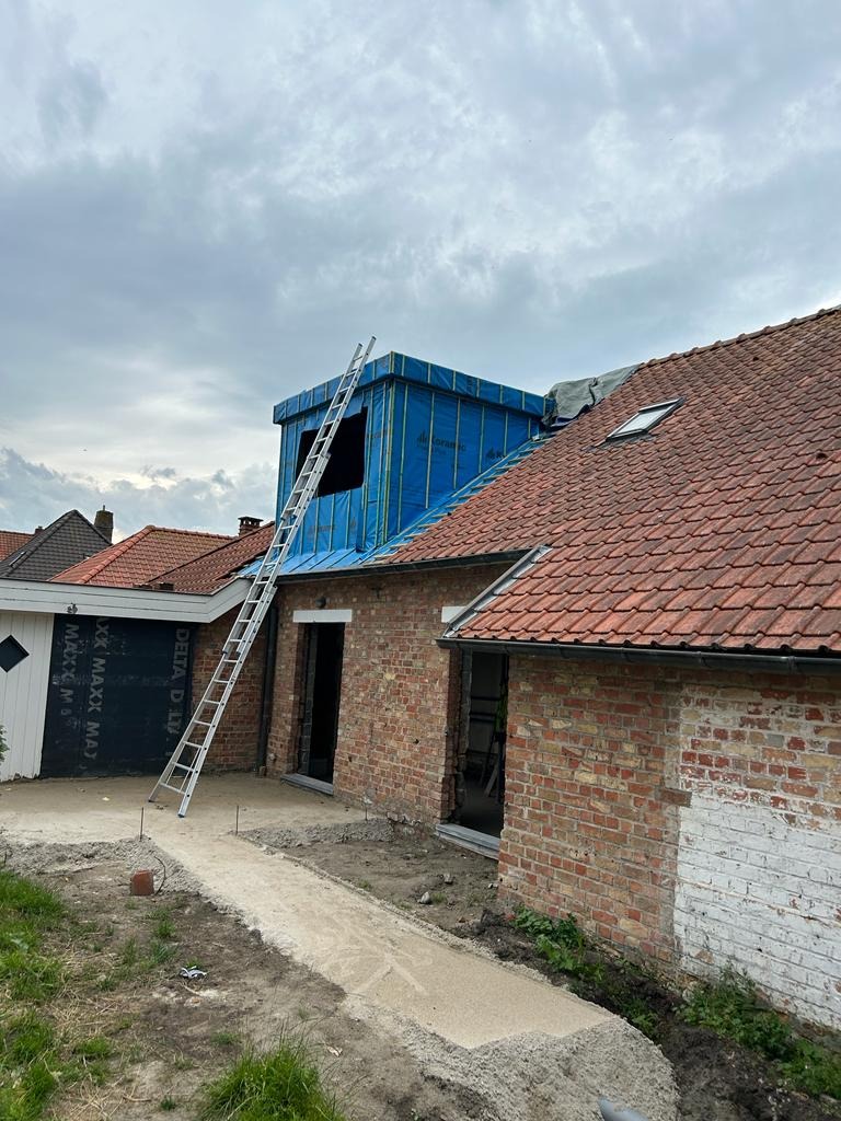 Budynek z cegły z nową nadbudówką pokrytą folią, drabina oparta o dach. Widoczne prace budowlane, przygotowanie do dalszych etapów wykończeniowych.