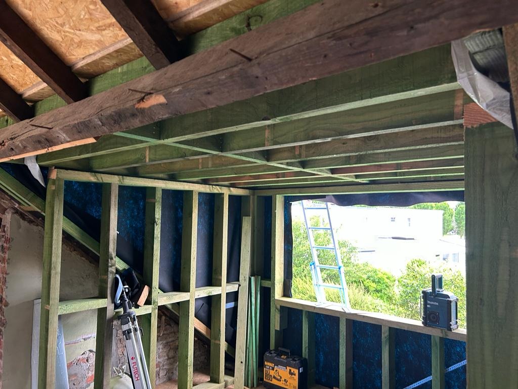 Wnętrze poddasza w trakcie adaptacji: drewniana konstrukcja szkieletowa pomalowana na zielono, widoczne narzędzia, drabina oparta o okno, surowe wykończenie ceglanej ściany.