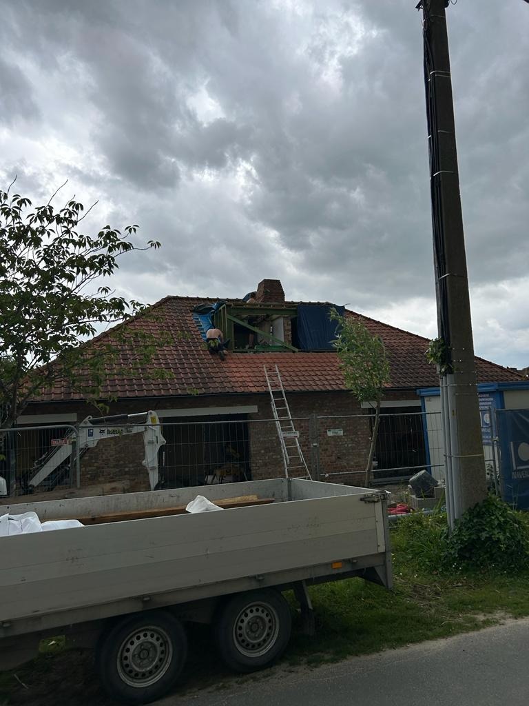 Demontaż uszkodzonej więźby dachowej z kominem na budynku z czerwonej cegły, widok z poziomu ulicy, z przyczepą i sprzętem budowlanym w kadrze.