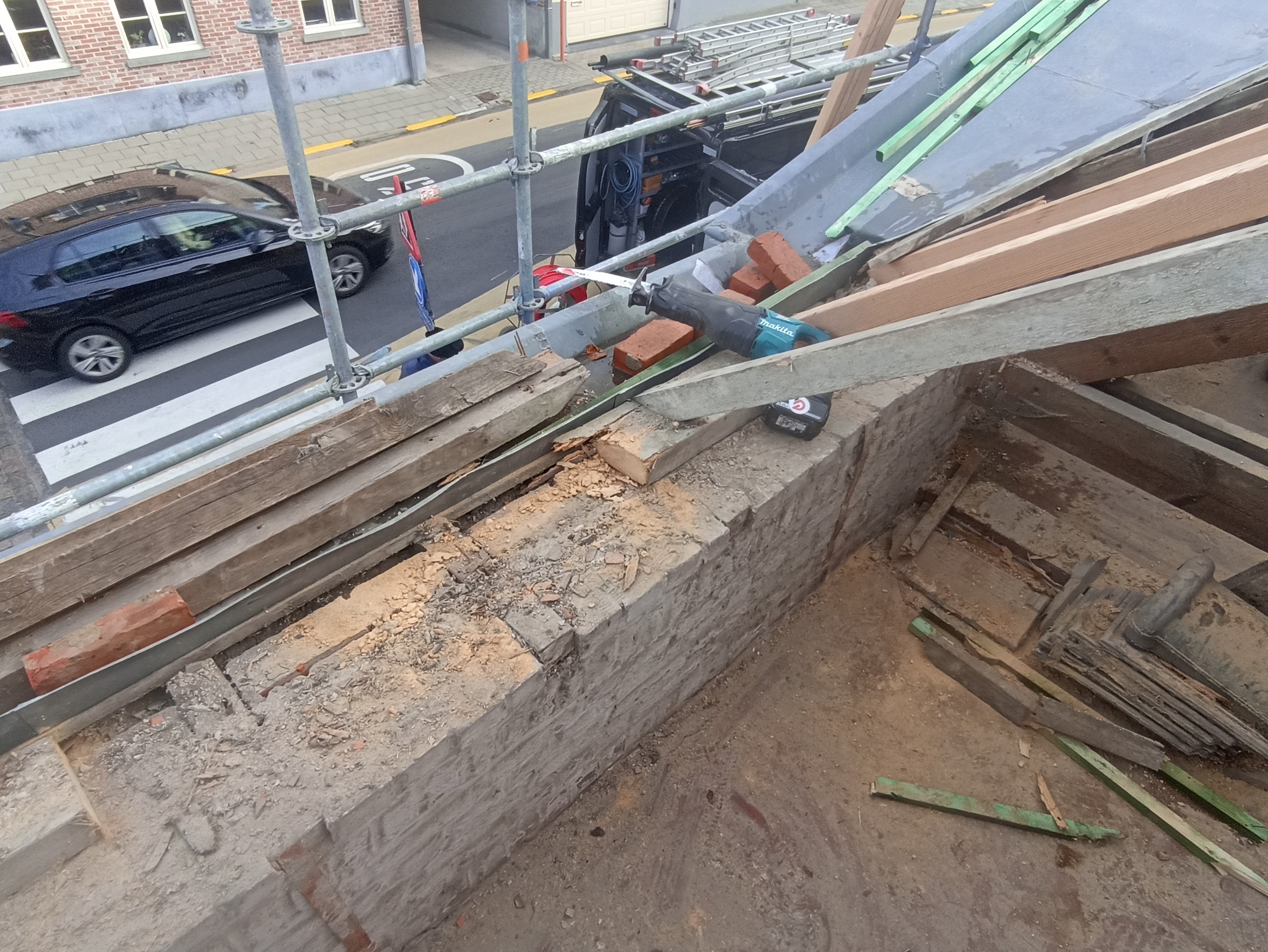 Demontaż dachu z użyciem piły szablastej Makita. Widoczne deski, cegły i rusztowanie. W tle ulica z przejściem dla pieszych i zaparkowany samochód.