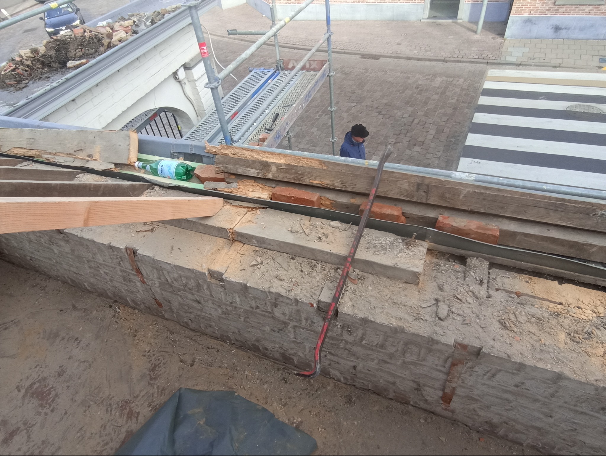 Demontaż uszkodzonych cegieł na dachu budynku w trakcie prac dekarskich. Widoczne rusztowanie, narzędzia i fragment ulicy z przejściem dla pieszych.