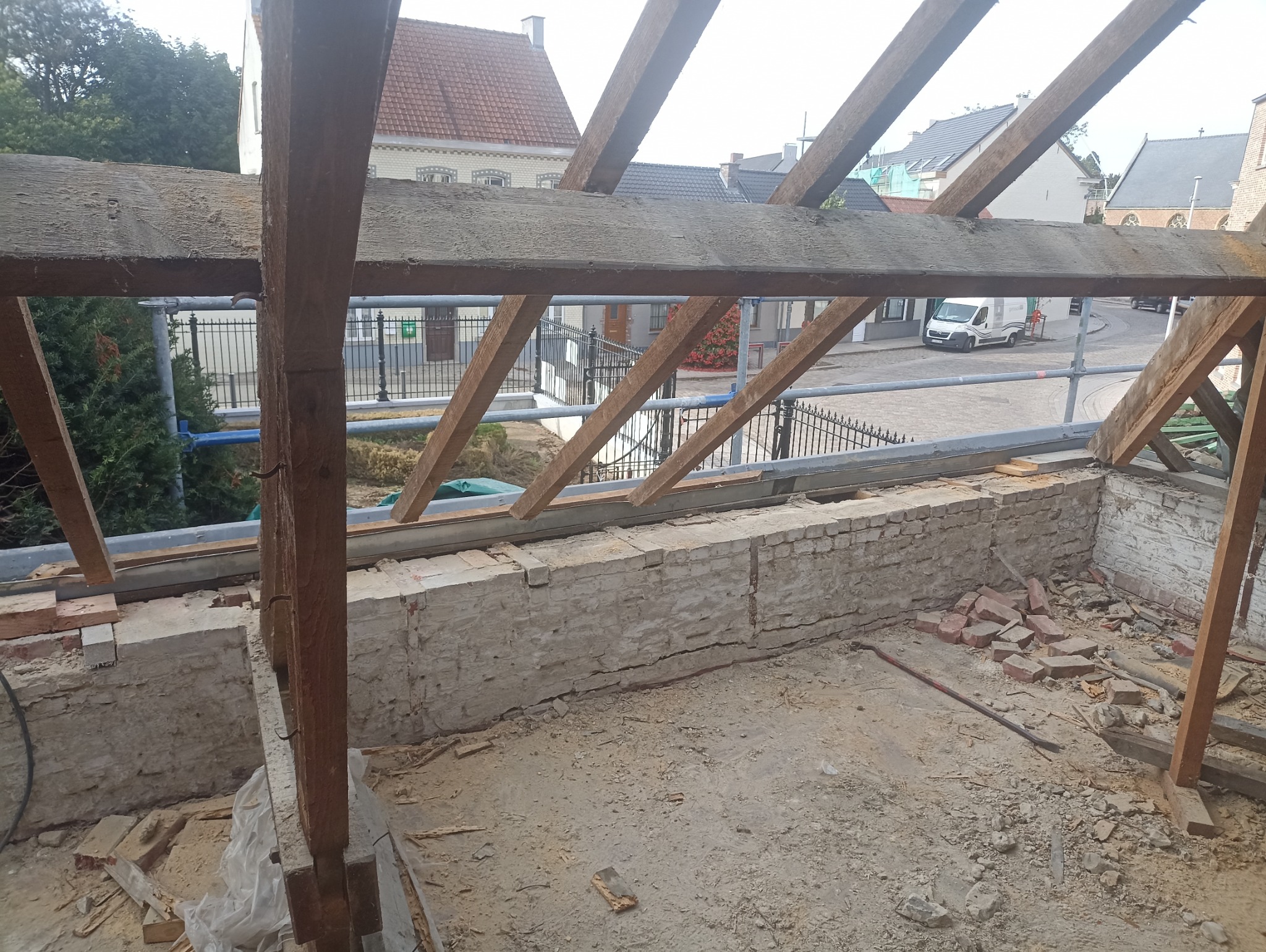 Wnętrze budynku w trakcie remontu dachu z widoczną więźbą dachową, fragmentami muru i porozrzucanymi cegłami. W tle widoczne budynki mieszkalne i fragment ulicy.