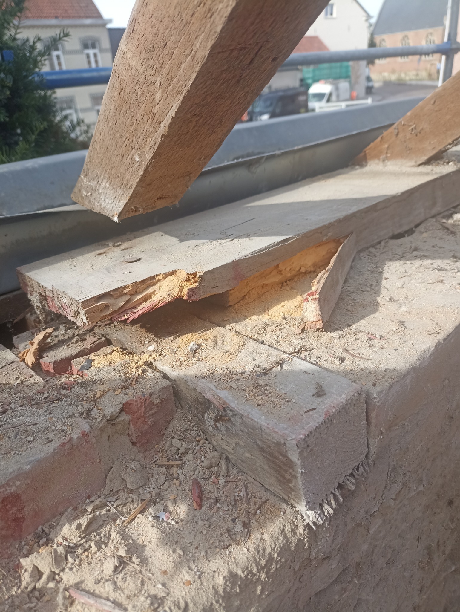 Fragment uszkodzonej więźby dachowej z widocznymi śladami zniszczeń, w tle zabudowa miejska. Drewno częściowo rozwarstwione, z ubytkami.