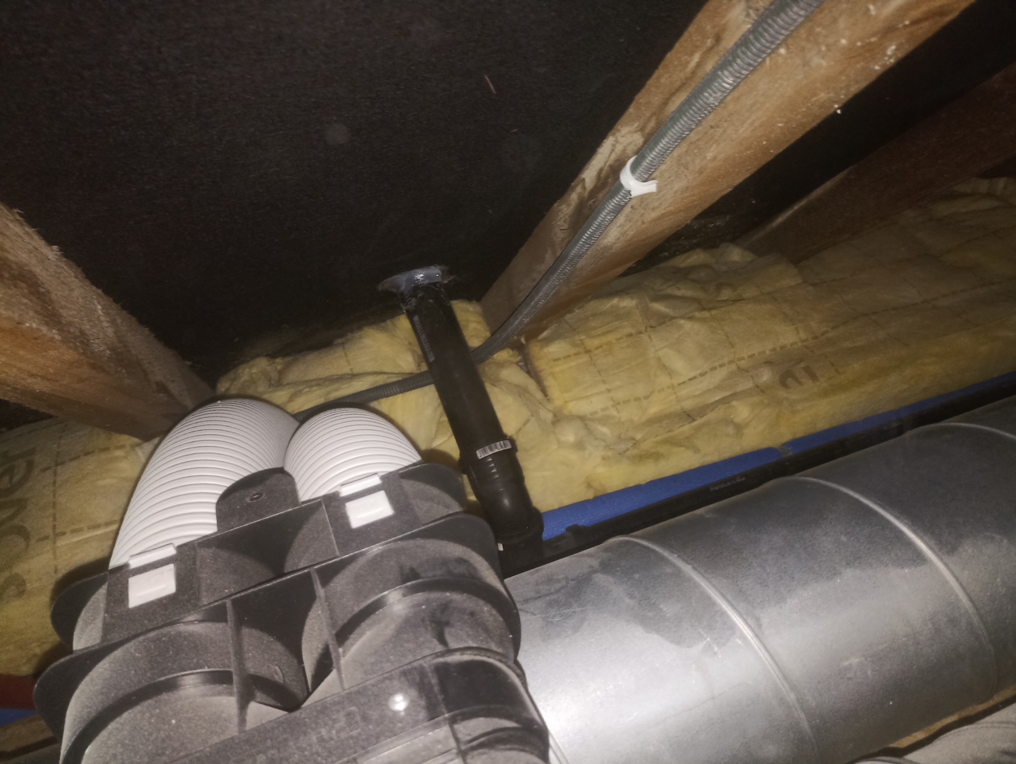 Żółta wełna mineralna między krokwiami, czarna rura wentylacyjna i białe, karbowane przewody w przestrzeni poddasza. Widoczne elementy instalacji wentylacyjnej.