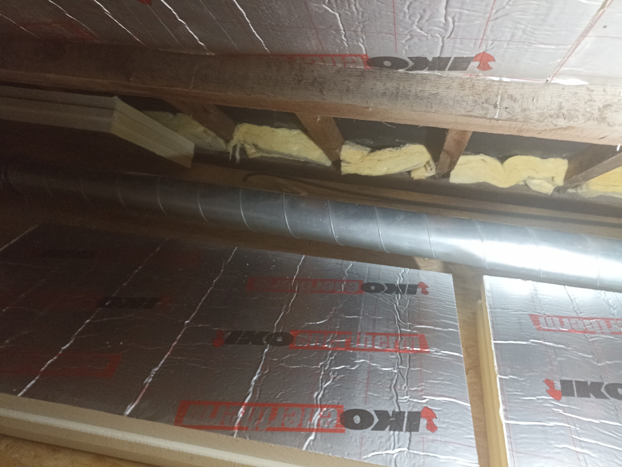 Ocieplone poddasze wełną mineralną i folią termoizolacyjną IKO, widoczne elementy drewnianej konstrukcji dachu i rura wentylacyjna.