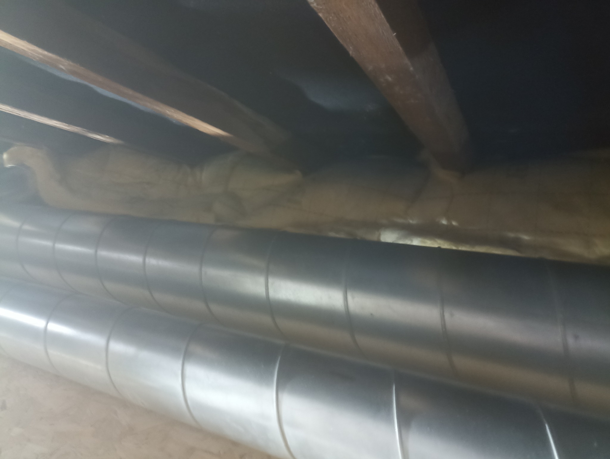 Dwa metalowe przewody wentylacyjne w przestrzeni poddasza, z widoczną izolacją między krokwiami. Niewyraźne oświetlenie podkreśla detale konstrukcji.