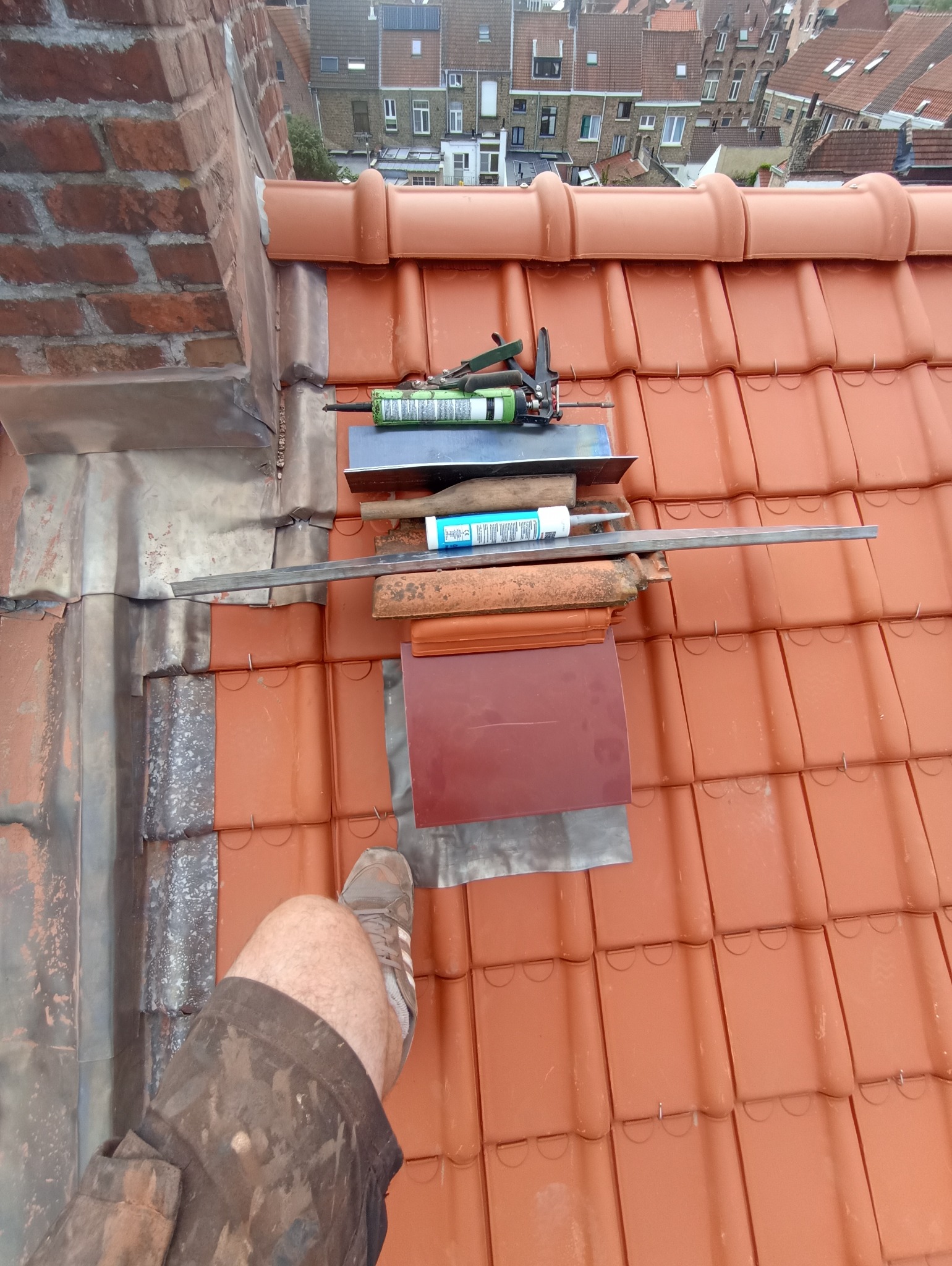 Czerwona dachówka ceramiczna z narzędziami dekarskimi i fragmentem nogi dekarza w ubrudzonych spodniach roboczych w rogu kadru. W tle zabudowa miejska.