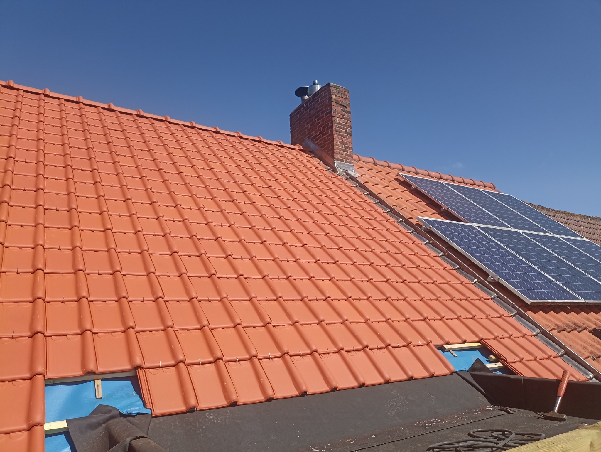 Czerwona dachówka ceramiczna na dachu domu, z kominem i panelami słonecznymi. Fragment dachu w trakcie układania, widoczne narzędzia i membrana dachowa.