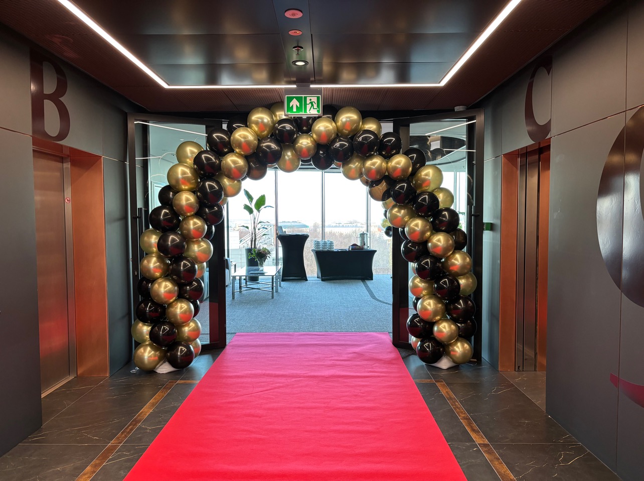 Wejście do biura zdobi łuk z czarno-złotych balonów i czerwony dywan, tworząc elegancką oprawę. W tle widoczne windy i przestrzeń biurowa.