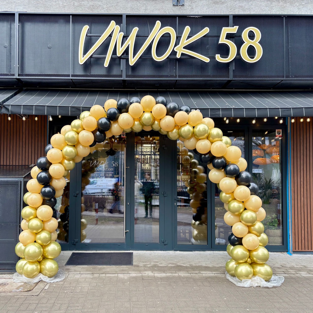 Dekoracja balonowa w odcieniach złota i czerni tworzy łuk nad wejściem do restauracji WOK 58, z widocznym menu na drzwiach i częściowo odbijającą się sylwetką osoby.