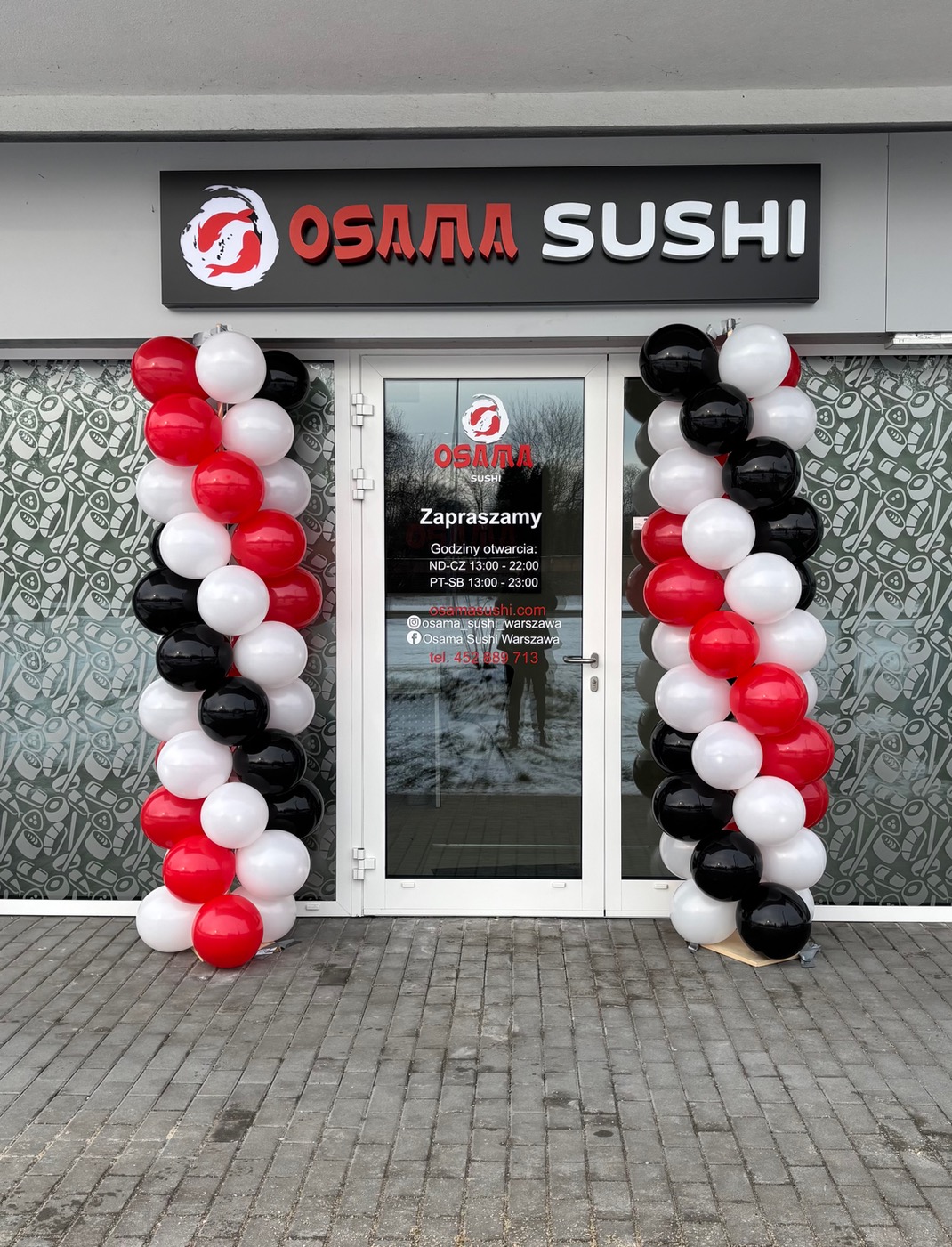 Wejście do Osamy Sushi udekorowane kolumnami z czerwono-biało-czarnych balonów. Nad drzwiami szyld z nazwą restauracji. Dekoracja eventowa przed lokalem gastronomicznym.