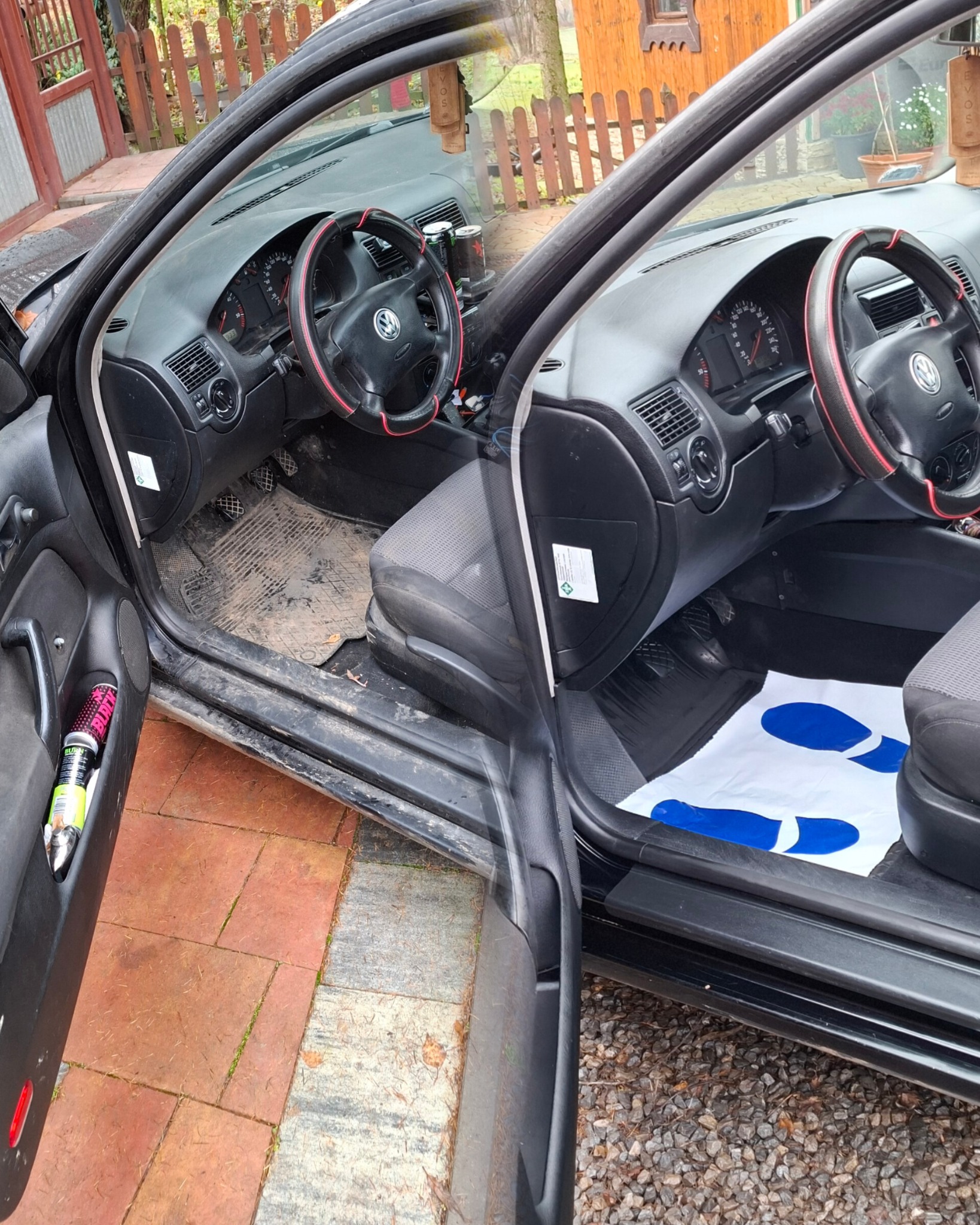 Wnętrze czarnego samochodu z otwartymi drzwiami, widoczne brudne dywaniki i deska rozdzielcza. Na podłodze leży papierowa mata z nadrukiem stóp.