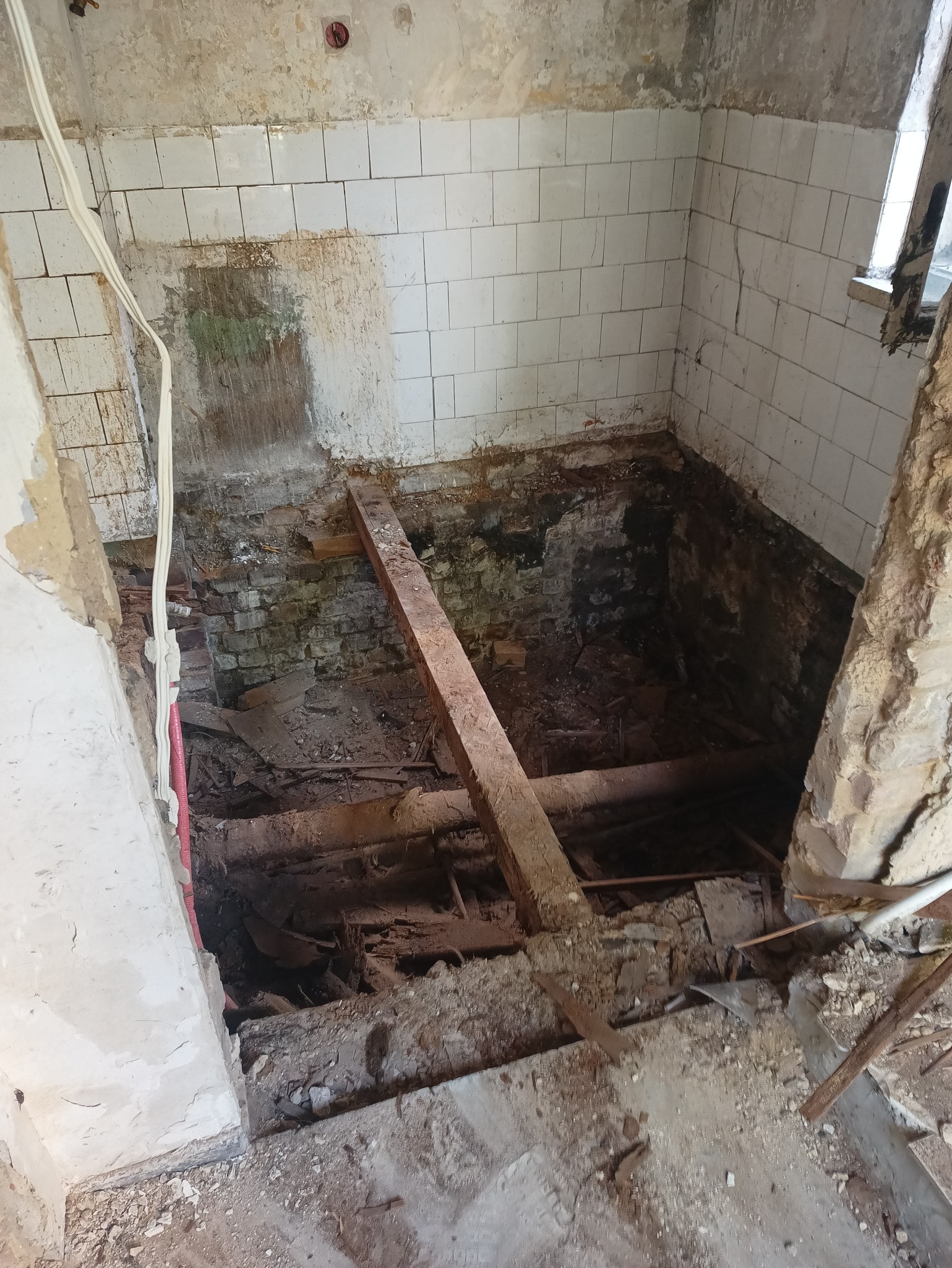 Zdemolowane pomieszczenie z odsłoniętą podłogą, fragmentami płytek na ścianach i wystającymi przewodami elektrycznymi, ukazujące stan przed remontem.