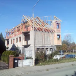 Budowa domu jednorodzinnego w stanie surowym otwartym z widoczną drewnianą konstrukcją dachu i elementami murowanymi, w słoneczny dzień.