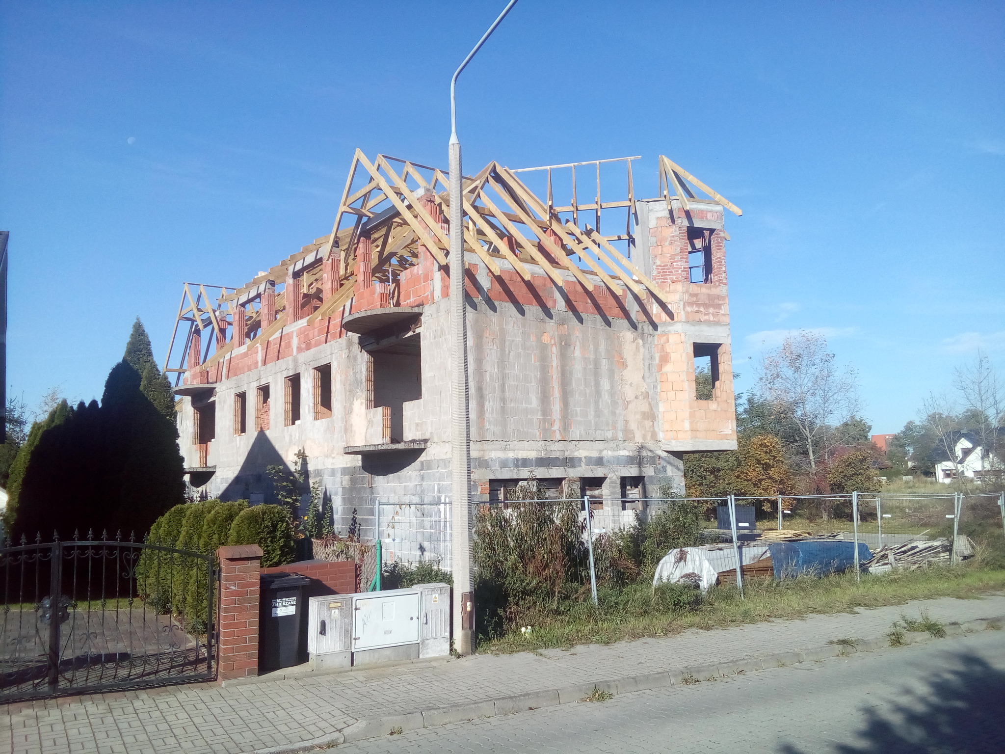 Budowa domu jednorodzinnego w stanie surowym otwartym z widoczną drewnianą konstrukcją dachu i elementami murowanymi, w słoneczny dzień.