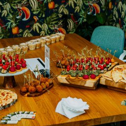 KERR FOOD sp. z o.o. - Stół cateringowy z przekąskami: koreczki z pomidorkami, kulki mięsne, pizza na drewnianej desce, sałatki w kubeczkach. W tle krzesła i ściana w tropikalne wzory.