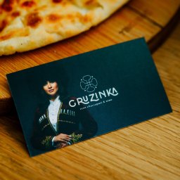 KERR FOOD sp. z o.o. - Karta wizytowa restauracji Gruzinka leży na drewnianym blacie obok kawałka chaczapuri. Karta prezentuje wizerunek kobiety w tradycyjnym stroju gruzińskim.