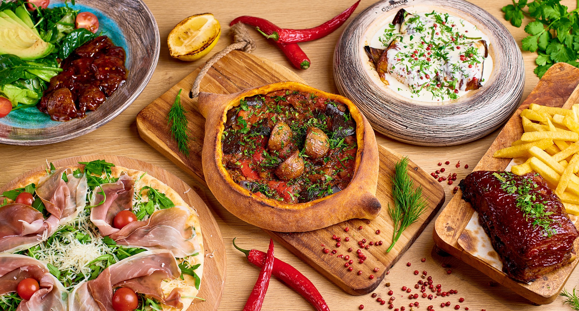 Bogaty stół z różnorodnymi daniami: chaczapuri, pizza z szynką parmeńską, żeberka BBQ, sałatka z awokado i bakłażan w sosie śmietanowym.